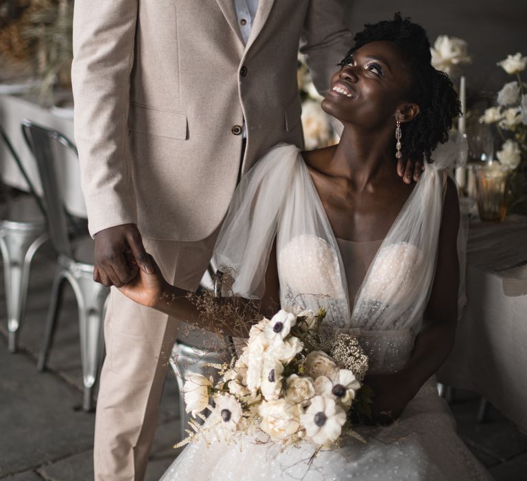 Groom in beige suit holding his brides hands whilst she sitting at the wedding reception table 