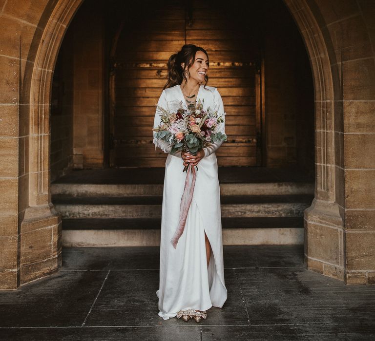 Bride stands in archway wearing wrap wedding dress carrying pastel bouquet 