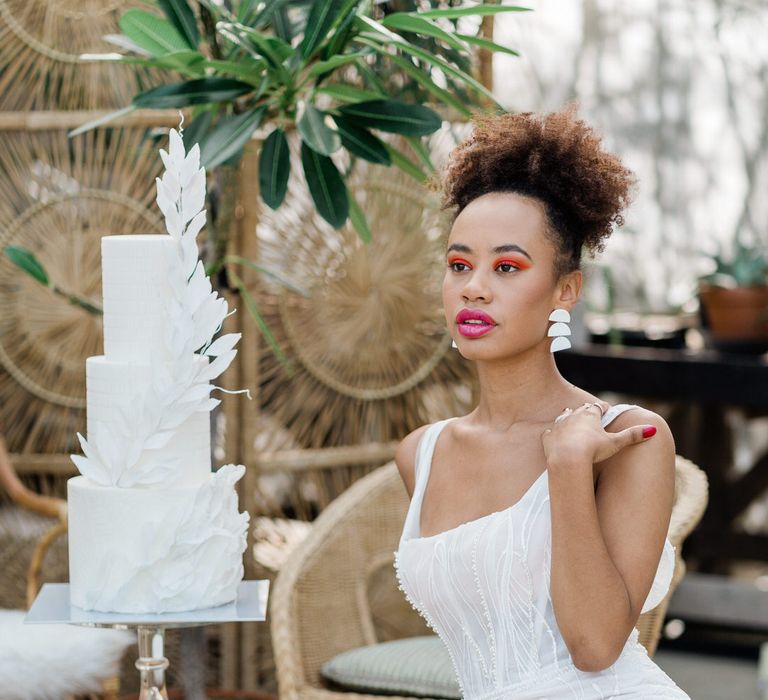 Bride in square neck wedding dress sat next to white three-tier textured wedding cake with leaf detail