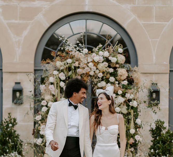 Stylish bride and groom in a white tuxedo and satin wedding dress and bow headdress holding hands whilst walking down their flower decorated aisle 