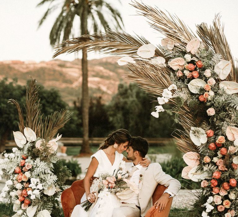 Boho bride and groom sitting on a velvet coral chair surrounded by pink roses, anthuriums ad dried grasses boho wedding flowers 