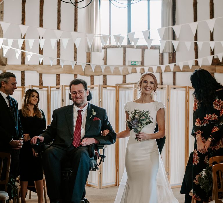 Bride in a fitted pronovnas wedding dress walking down the aisle with her dad in a wheelchair 