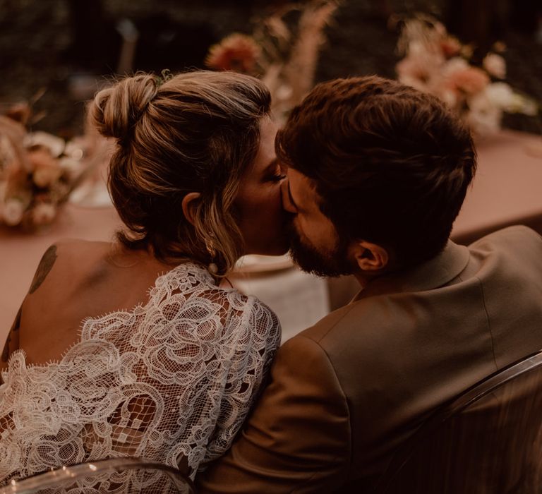 Groom in a brown suit kissing his bride in a one shoulder lace wedding dress at their intimate forest reception 