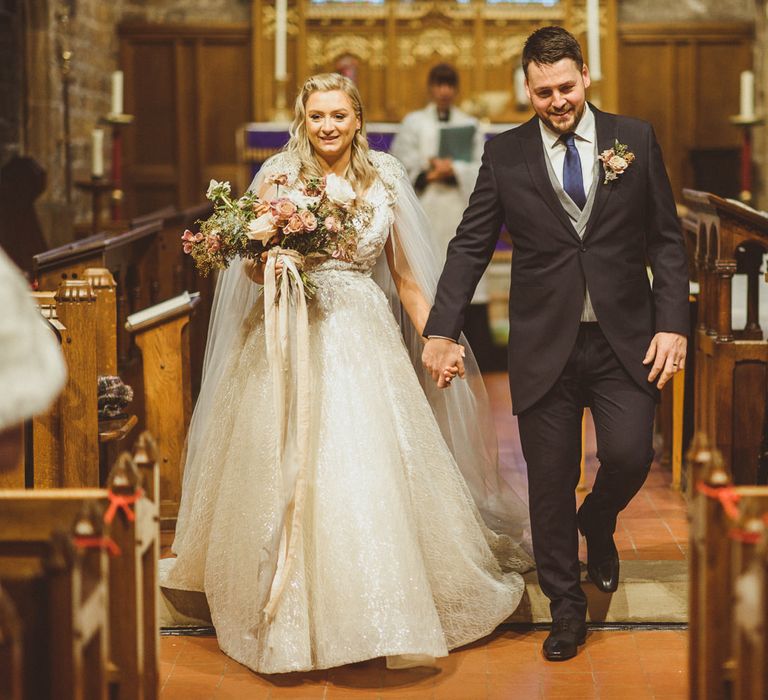 Groom in a navy morning suit holding his brides hand in a princess wedding dress walking up the aisle as husband and wife 