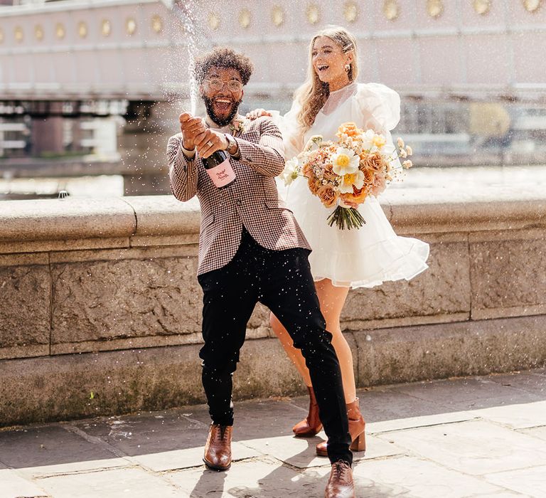 Groom in black trousers and patterned blazer spraying champagne on Albert's Bridge in Chelsea