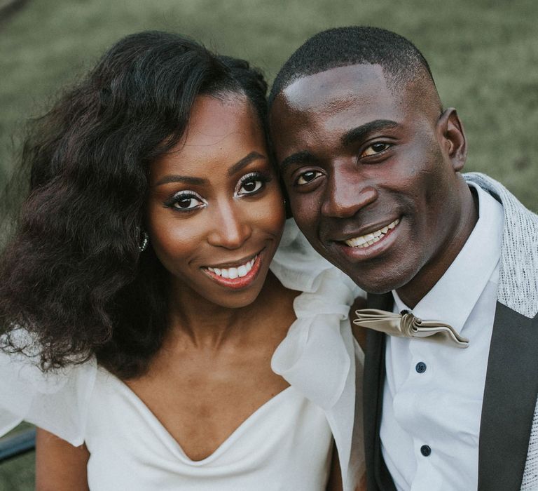 Smiling bride with short curled hair in white satin dress with shoulder bows and white and green bouquet sits on a bench with smiling groom in grey suit jacket and bow ties