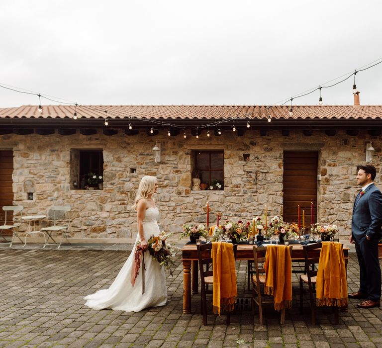 The bride and groom standing at each end of their outdoor wedding table, smiling at each other