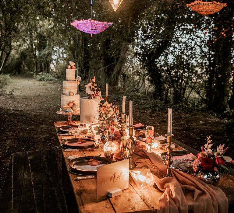 Wooden wedding tablescape with pink table runner, silver candles and tropical floral decor with suspended umbrellas and hanging lights for festival themed wedding