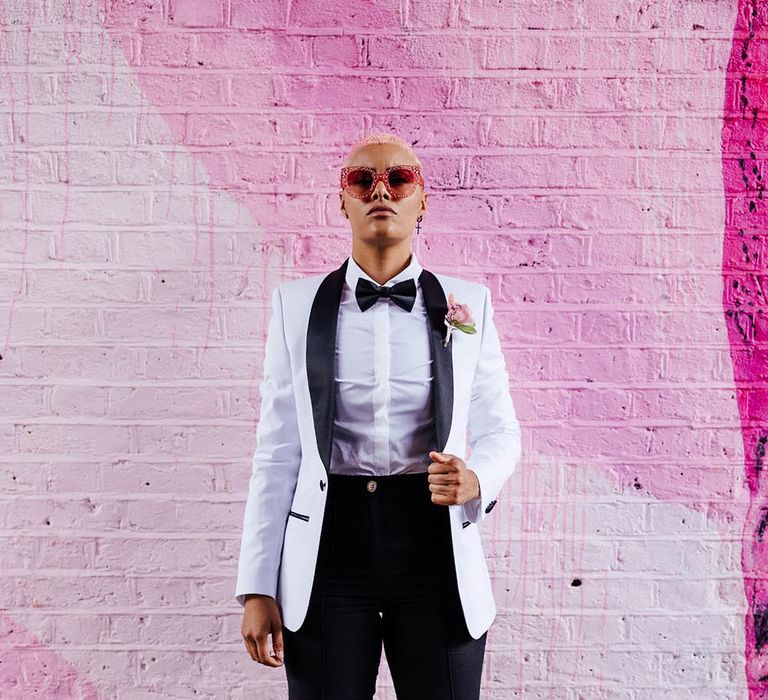 Bride in a white tuxedo jacket with black bow tie standing in front of a pink wall 