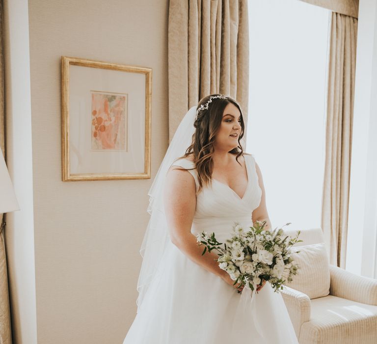 Bride stands whilst holding white floral bouquet and has her dark hair in loose curls with delicate floral headband reaching her veil