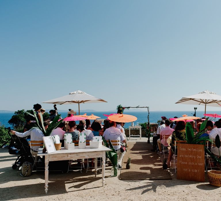 Outdoor wedding ceremony in France with guests shielding from the sun under orange and pink parasols 