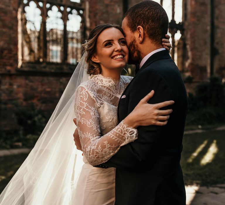 Beautiful bride in a lace wedding dress and cathedral length veil smiling as she embraces her husband in a black suit 