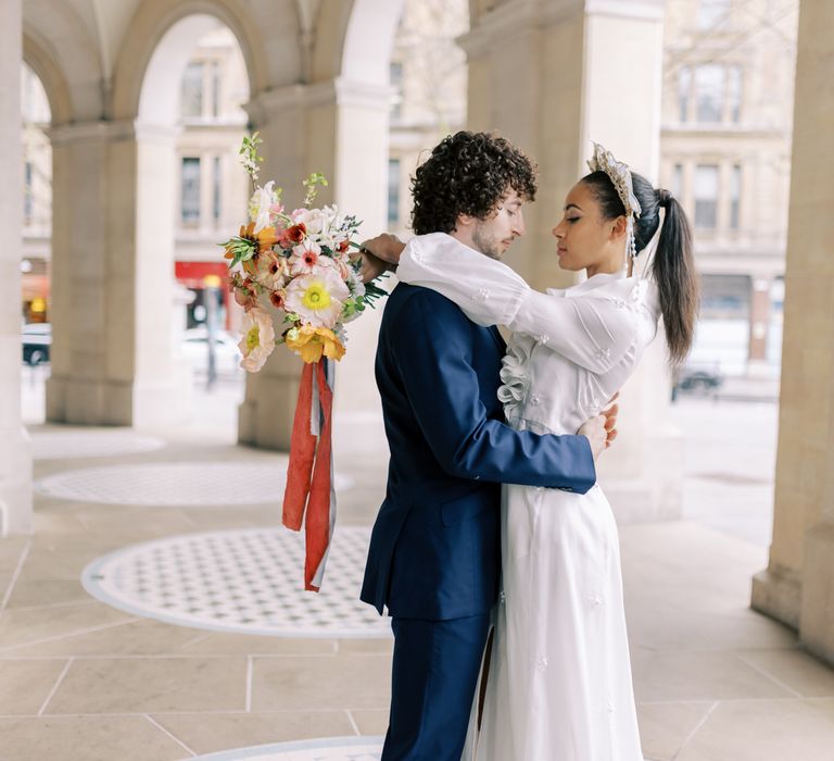 Bride & groom embrace outdoors as bride holds floral bouquet tied with ribbon