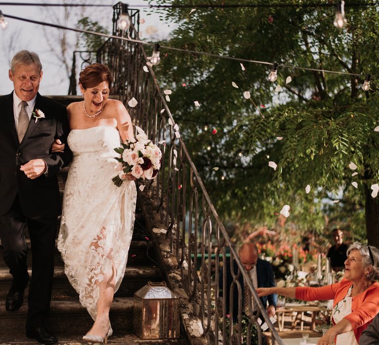 Bride & groom walk down staircase whilst wedding party throw confetti and fairy lights twinkle in the trees