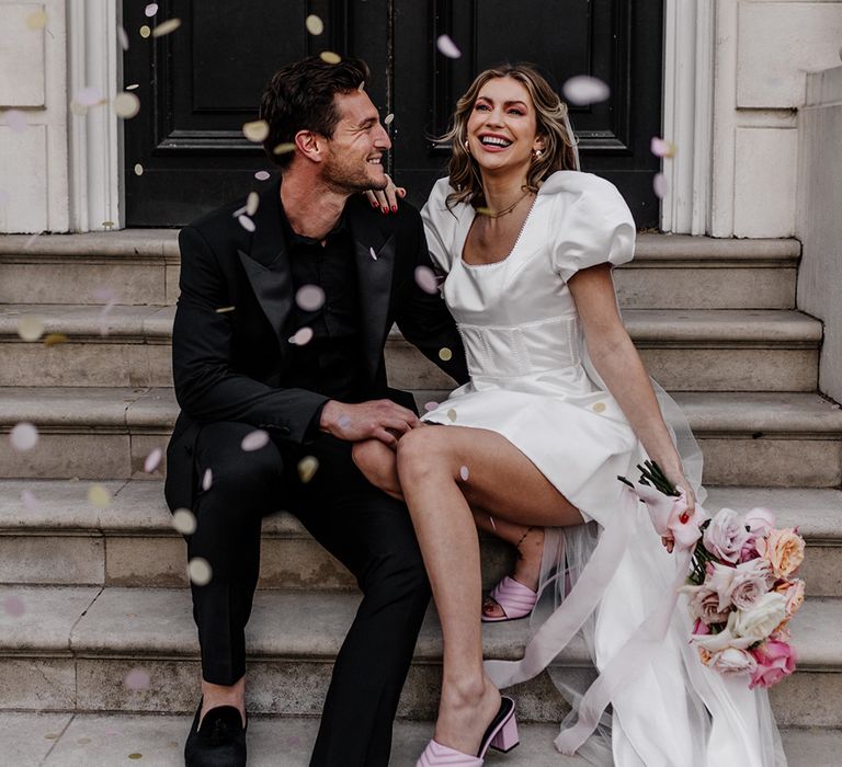 Modern luxury wedding day moment with bride and groom sitting on some steps as confetti is thrown over them