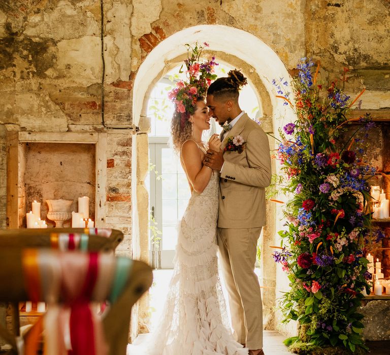 Bride in a layered ruffle skirt wedding dress and colourful flower crown standing at the altar with her husband in a beige suit next to a vertical flower arrangement 