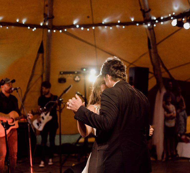 Groom in dark brown suit dances with bride in white Elbeth Gillis wedding dress inside tipi wedding reception at home farm wedding