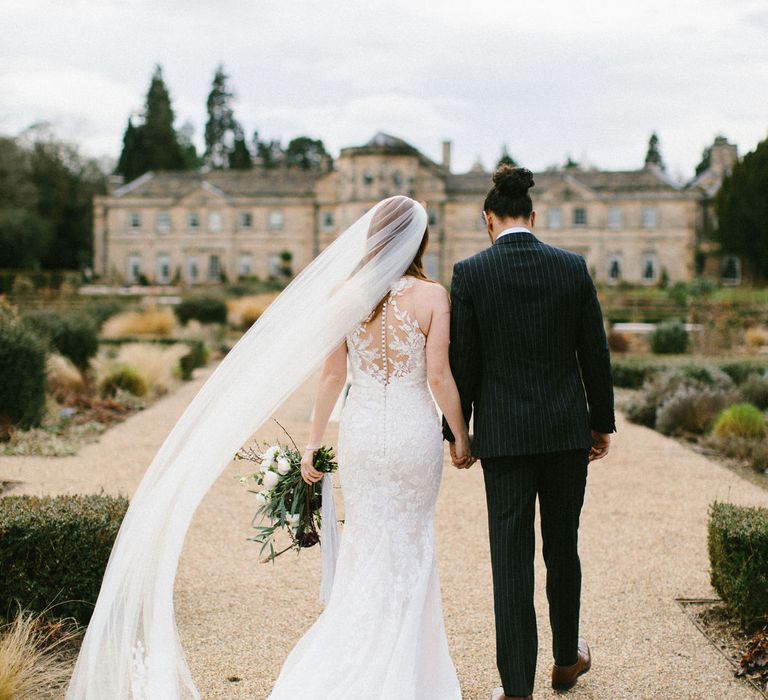 Pride in a lace Pronovias wedding dress and long veil holding hands with her groom in a pinstripe wedding suit in from of their wedding venue 