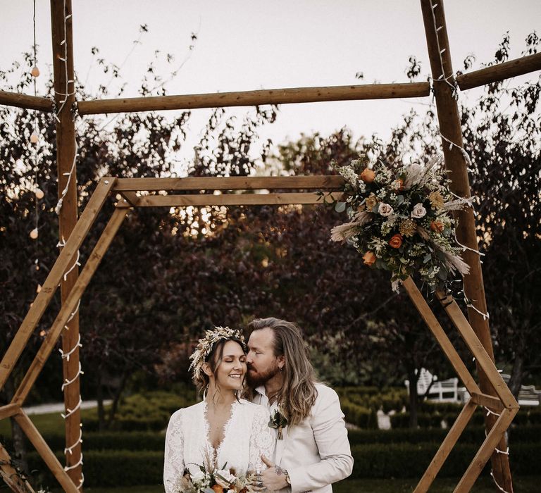 Groom in a beige suit with long hair kissing his bride's cheek in front of their hexagonal wooden frame altar 