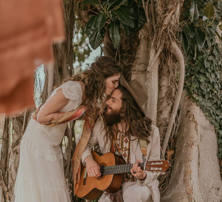 Bride kisses groom as he plays guitar under ficus tree as bride wears boho chic wedding gown and groom wears suede hat 