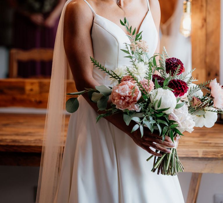 Smiling bride in white cami wedding dress with shoulder veil detailing looks over her shoulder as she holds white, pink and green bridal bouquet before ceremony at The West Mill Derby