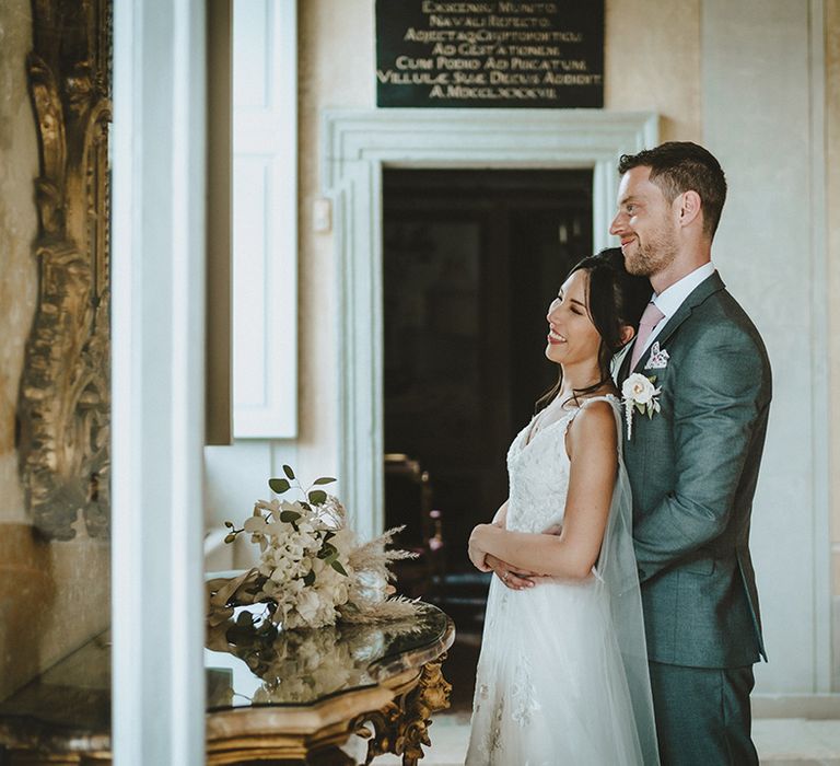 Groom holds his arms around bride as they look out the window on their wedding day