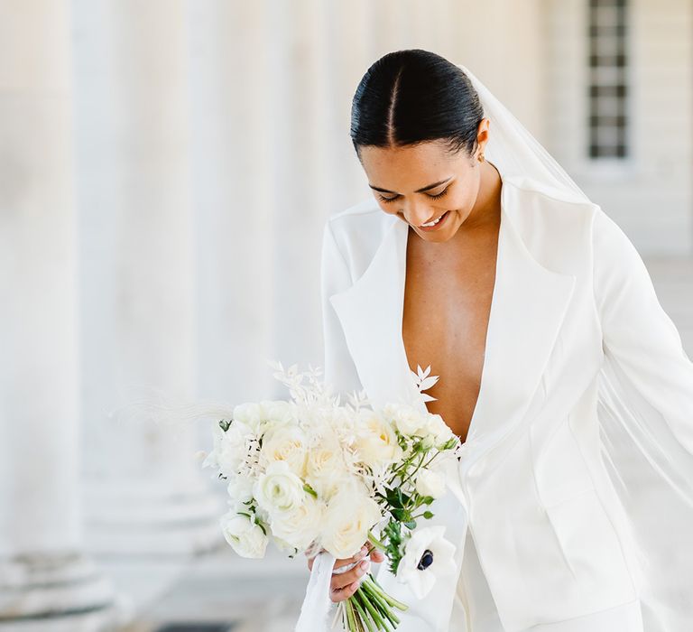 A bride wears a low cut white suit and holds a bouquet filled with white flowers and anemones. Her brown hair is parted in the middle and tied low. She has small gold hooped earrings and a veil.