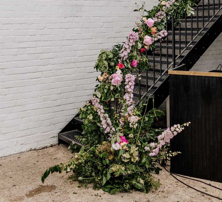 Metal stairs in courtyard wrapped with green foliage and pink, purple and. orange flowers for wedding at Loft Studios London