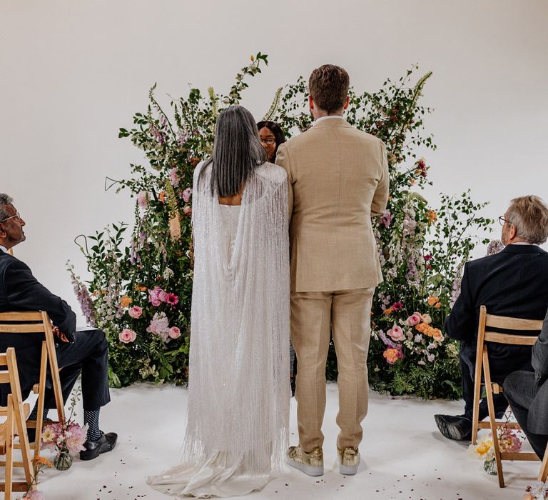 Bride in satin Halfpenny London wedding dress and tasselled bridal cape stands with groom in linen suit and gold Nike Air Force 1 trainers during wedding ceremony at Loft Studios London