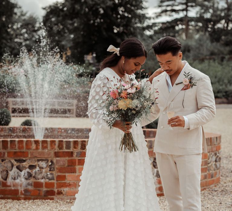 Bride in white long mesh sleeve spotted Vagabond wedding dress and bridal hair bow holding mixed bridal bouquet stands holding shoulder of groom in white shawl collared shirt and double breasted linen suit with brown shoes next to fountain outside in gardens at Wasing Park wedding