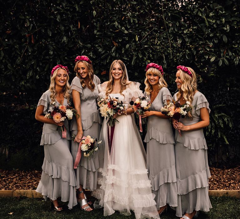 Bridesmaid in white Halfpenny London Mayfair dress holding mixed bridal bouquet stands in the middle of bridesmaids in grey ruffle bridesmaid dresses and pink headbands before church ceremony in Cornwall 