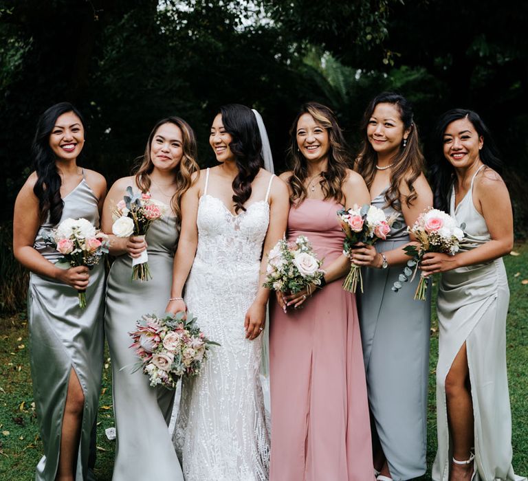 The Bridal party smile in blush pink and sage green satin bridesmaid dresses as bride sparkles in outdoor setting