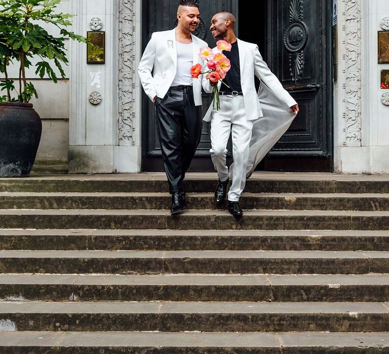 Two grooms at city elopement in leather trousers, a white suit with cape and a red flower bouquet 