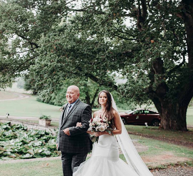 Bride wears fitted long dress with chiffon puff detail at the bottom holds arms with her father in checked blue suit at Cefn Tilla Court 