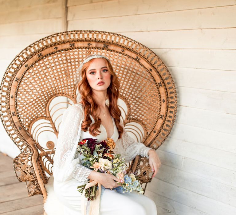 Bride in long lace sleeve wedding dress and Juliet cap wedding veil holding colourful bridal bouquet sits in large rattan chair on wooden veranda