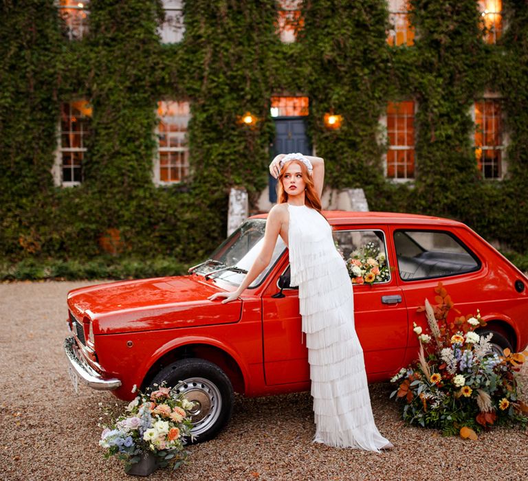 Bride in white headband and long fringed wedding dress leans on small red car filled with florals outside manor house covered in ivy