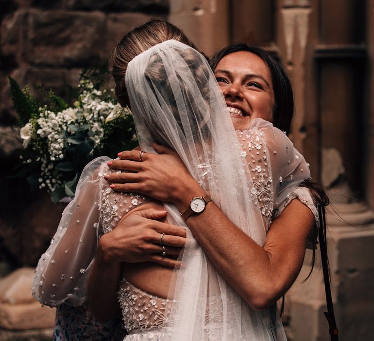 Bride in open back Julie Vino embroidered wedding dress and veil hugs smiling wedding guest after church wedding ceremony