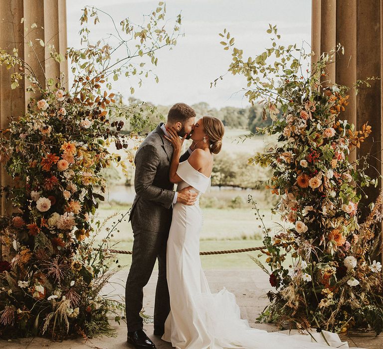 Bride & groom kiss in front of floral archway on their wedding day