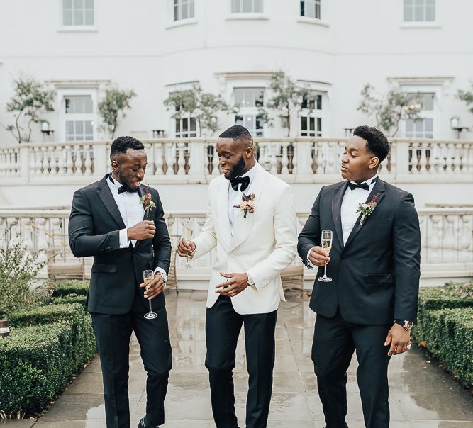 Groom wears white jacket for black tie wedding whilst he stands with his groomsmen who wear black tie and hold glasses of champagne