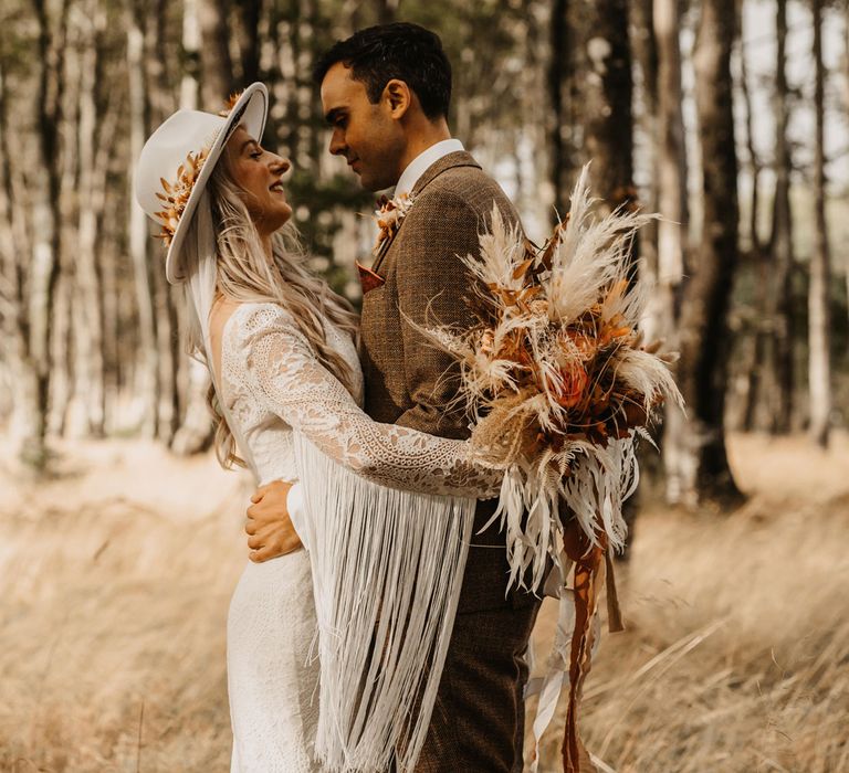 Bride in boho tassel two piece wedding dress and hate holding white and orange boho bouquet hugs groom in brown tweed suit as they stand in woodland