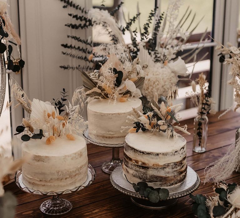 Three naked wedding cakes complete with white frosting next to dried florals