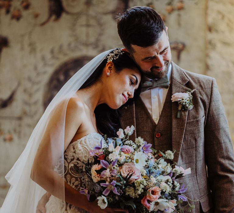 Bride in lace and tulle wedding dress and veil holding pink, purple and blue bridal bouquet rests head on the shoulder of groom in brown check tweed suit during wedding ceremony