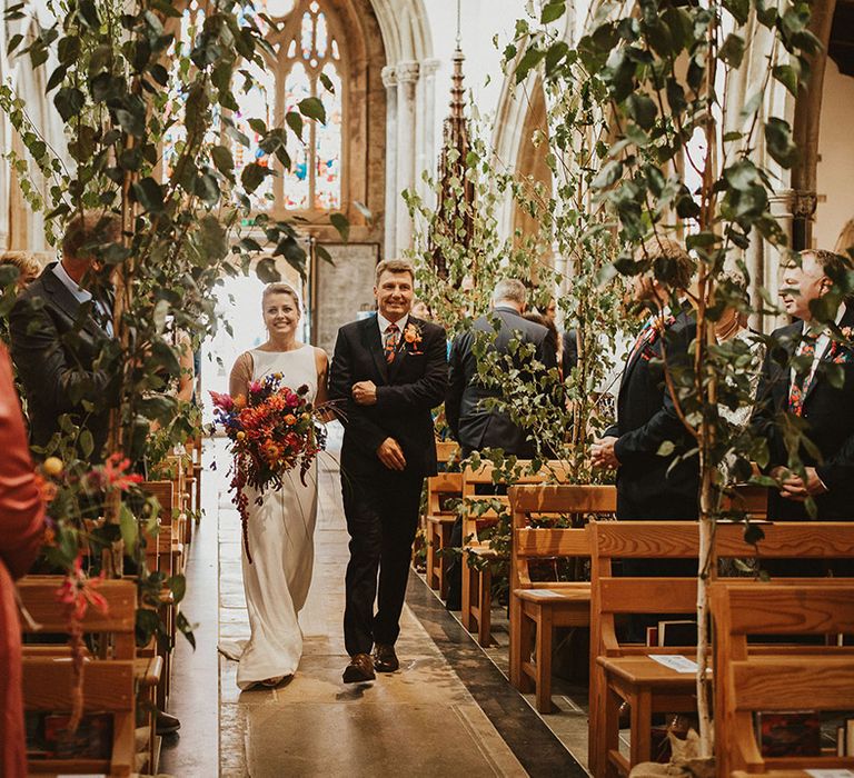 Father of the bride walking his daughter down the church aisle decorated with trees 