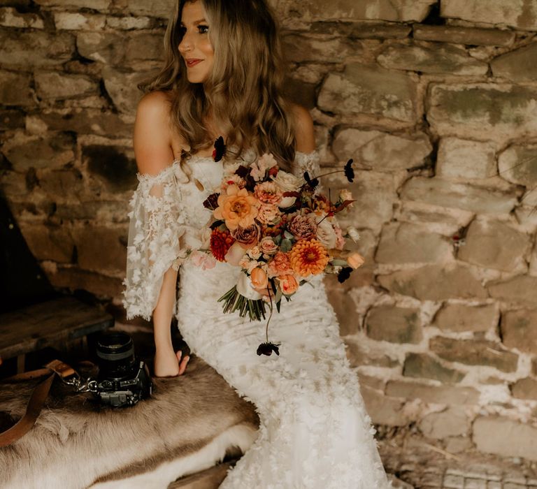 Bride in off the shoulder wedding dress with 3D applique flowers sits on fur covered bench holding mixed peach bridal bouquet 