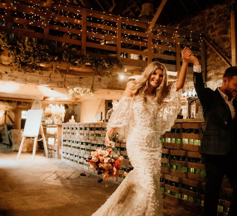 Bride in boho wedding dress with 3d applique florals holding mixed autumnal bridal bouquet walks into rustic wedding breakfast in stone barn holding hands with groom in dark suit