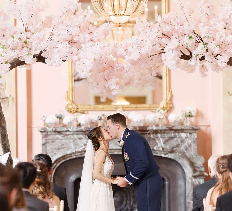 Bride & groom kiss under blossom archway on their wedding day during ceremony