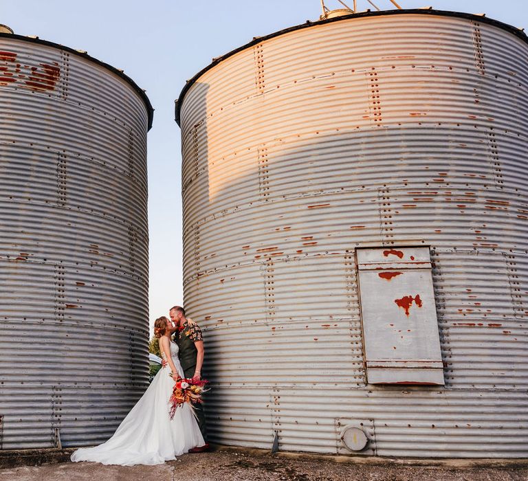 Bride in corset back wedding dress holding mixed rustic bouquet stands with groom in short sleeve patterned shirt by large grain store at farm festival wedding