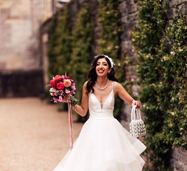 Bride in wedding dress with layered tulle skirt, feather headband and pearl necklace holds beaded bag and pink, red and white mixed bridal bouquet