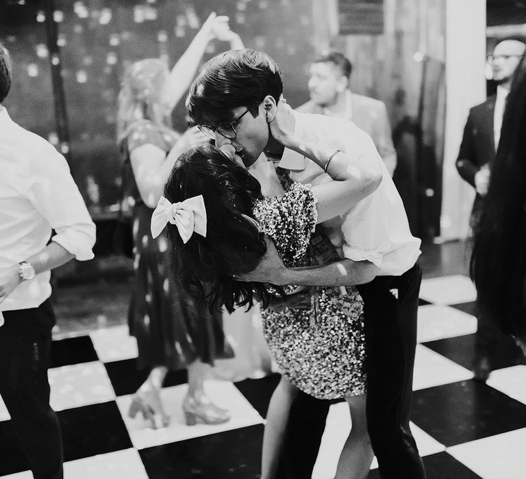 Bride with bow and short pink dress kisses groom in white shirt on dance floor during evening wedding party