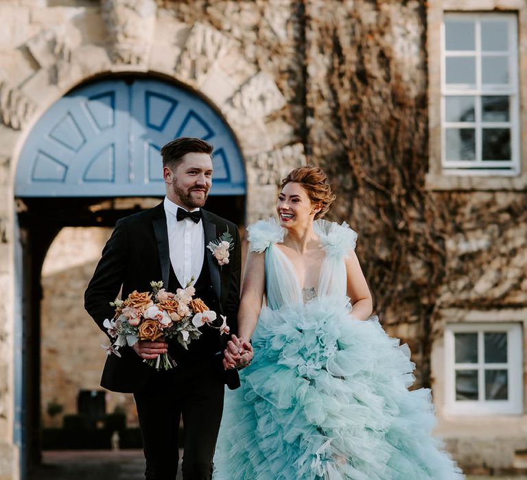 Bride and groom holding hands at their black tie wedding with groom in a tuxedo and horseshoe waistcoat and bride in a blue ruffle wedding dress 